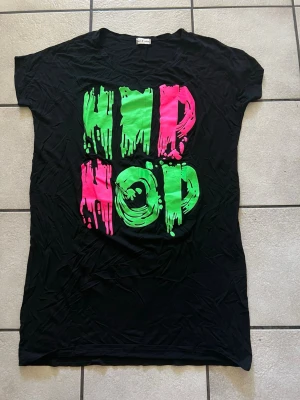 Svart t-shirt med neon HIP HOP-tryck storlek M/L - Cool svart t-shirt med stort färgstarkt HIP HOP-tryck i neonrosa och neongrönt framtill. T-shirten har korta ärmar och en avslappnad passform. Perfekt för dig som vill sticka ut med streetstyle-vibe.