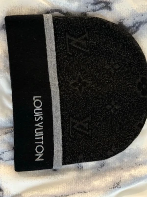 Svart mössa  - Snygg svart mössa med diskret monogrammönster och bred grå kant. Märkets logga är broderad i vitt på kanten. Mössan är mjuk och stretchig, perfekt för kalla dagar och ger en exklusiv touch till din outfit.