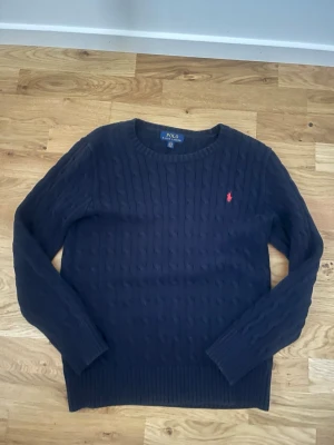 Mörkblå kabelstickad tröja Polo Ralph Lauren - Kabelstickad mörkblå tröja från Polo Ralph Lauren med rund halsringning och röd broderad logga på bröstet. Tröjan har långa ärmar och ribbade muddar vid ärmslut och nederkant. Perfekt för dig som gillar klassisk och stilren look.