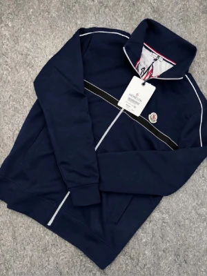Marinblå ziptröja från Moncler - Snygg marinblå ziptröja från Moncler med vit piping och svart rand över bröstet. Tröjan har hög krage, dragkedja framtill och ett stort Moncler-tryck med bergsmotiv på insidan. Perfekt för dig som gillar streetwear och exklusiva detaljer. Taggen borta annars ny skick