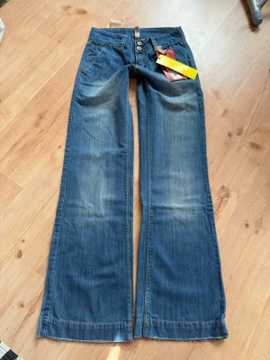 NYA Lowwaist, vida jeans Vero Moda - Säljer ett par snygga blå bootcut jeans med dubbla knappar i midjan och klassiska fem fickor. Jeansen har en lätt tvättad look och vida ben. Perfekta för dig som gillar retrostil och vill ha ett par jeans som sticker ut. midja 76, innerbenslängd 86! Budas ut på live 19.00 idag ! 