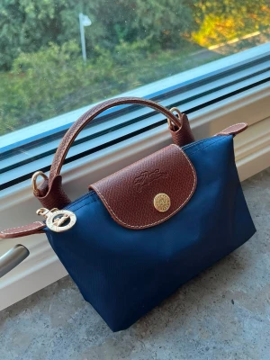 Mörkblå handväska med bruna detaljer - Snygg mörkblå handväska f med bruna läderhandtag och lock. Väskan har guldfärgade metalldetaljer och klassisk logotyp på locket.  Perfekt storlek för det viktigaste.