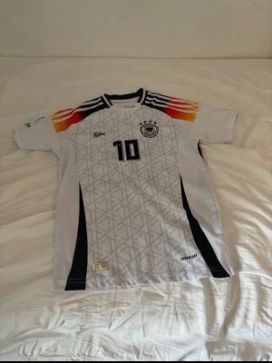 Tysklands fotbollströja Musiala #10 - Säljer en vit Tyskland fotbollströja med nummer 10 och namnet Musiala på ryggen. Tröjan har svarta detaljer, geometriskt mönster framtill och ränder i svart, rött och gult på axlarna. Perfekt för dig som älskar fotboll och vill stötta landslaget.