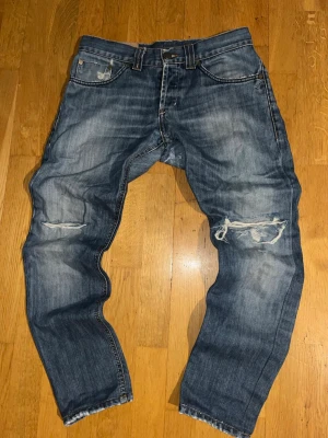 Dondup jeans  - Säljer ett par blå jeans med tydliga slitningar och hål på båda knäna. Jeansen har en klassisk femficksmodell och raka ben. Materialet är denim i bomull och de har en cool, använd look som passar streetwear-stilen.