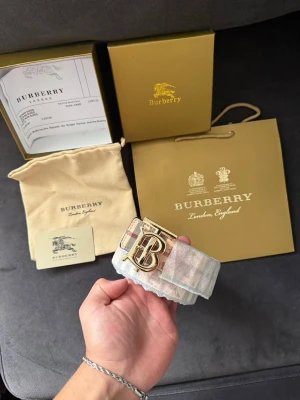 Burberry bälte - Säljer ett tvåsidigt Burberry bälte med klassiskt rutigt mönster i beige, brunt och rött. svart design på andra sidan. Perfekt accessoar för att lyfta din outfit.