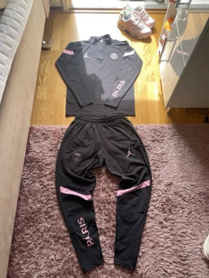 Svart PSG träningsset med rosa detaljer - Säljer ett svart PSG träningsset från Jordan med rosa detaljer. Setet består av en långärmad tröja med halv dragkedja och matchande byxor. Både tröjan och byxorna har PSG-logga och Jordan-logga samt texten PARIS i rosa. Perfekt för träning eller chill.