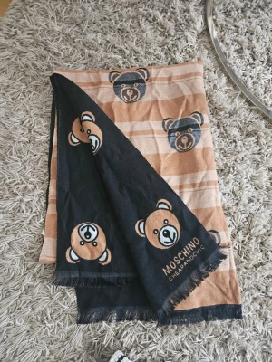 Moschino sjal med björnmönster - Säljer en snygg sjal från Moschino med ikoniskt björnmönster i beige, svart och vitt. Sjalen har fransade kanter och är tillverkad i ett mjukt tyg som känns skönt mot huden. Perfekt accessoar för att lyfta din outfit med en lekfull touch.
