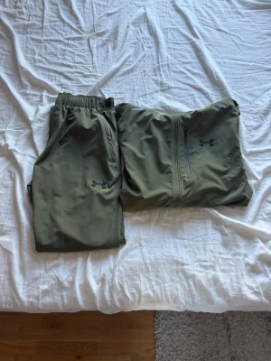 Olivgrön träningsset från Under Armour - Säljer ett olivgrönt träningsset från Under Armour med matchande byxor och jacka. Jackan har huva, dragkedja och fickor, samt logga på bröstet. Byxorna har resår i midjan och logga på benet. Perfekt för utomhusträning eller löpning.