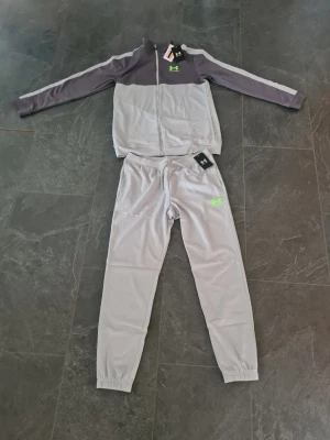  träningsset från Under Armour - Snyggt träningsset från Under Armour i ljusgrått med mörkgrå ärmar och detaljer. Setet består av en zip-jacka och matchande byxor med resår i midja och benslut. Under Armour-logga i neon på både jacka och byxa. Perfekt för träning eller chill.