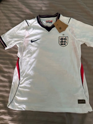 England Nike fotbollströja vit M - Säljer en vit England fotbollströja från Nike med broderad landslagssköld och svart Nike-logga. Tröjan har svarta och röda detaljer, V-ringad krage med mörkblå kant och texten 'HAPPY AND GLORIOUS' i nacken. Tillverkad i lätt och ventilerande material.