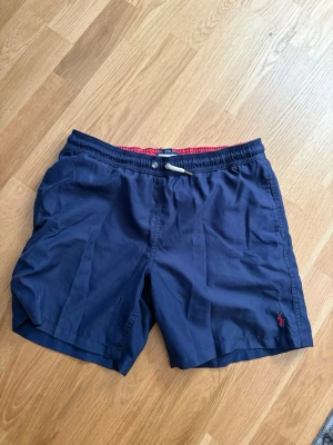 Polo Ralph Lauren badbyxor  - Snygga marinblå shorts med elastisk midja och snörning. Shortsen har en röd broderad logga på benet och röd insida i midjan. Perfekta för sommardagar och har en klassisk, avslappnad passform.