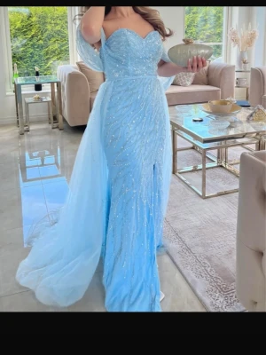 Ljusblå långklänning med glitter - Magisk ljusblå långklänning med glittriga detaljer och offshoulder-design. Klänningen har en elegant slits framtill och ett svepande släp i tunn tyll. Perfekt för dig som vill sticka ut på bal eller fest.