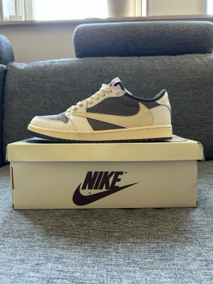 Nike x Travis Scott grå/beige sneakers - Nike x Travis Scott sneakers i grått och beige med omvänd swoosh på sidan. Lågt skaft, vita snören och detaljer i rött på plösen. Ovandelen är i mocka och läder, med perforerad tåbox för extra ventilation. Snygg och unik design som sticker ut.