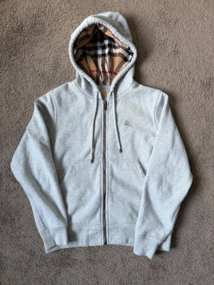 Burberry zip hoodie  - Snygg ljusgrå hoodie med dragkedja och kängurufickor. Huvan har ett klassiskt rutigt foder i beige, svart, vitt och rött. Tröjan har ribbade muddar vid ärmslut och nederkant samt justerbara snören i huvan. Perfekt för en chill och avslappnad stil. Pris kan diskuteras privat mvh ☺️