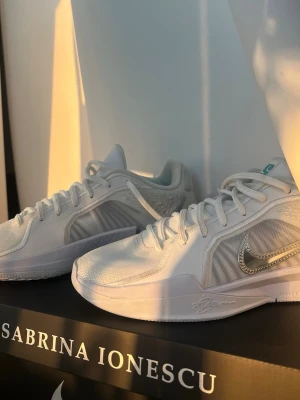 Nike Sabrina Ionescu vita sneakers - Snygga vita Nike Sabrina Ionescu basketskor med meshpaneler och silvrig Swoosh på sidan. Skorna har lågt skaft, vita snören och detaljer i ljusgrått. Perfekta för dig som gillar stilrena och sportiga sneakers med modern design.