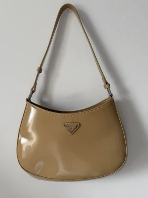 Beige axelremsväska i lack  - Snygg beige axelremsväska i glansigt lackmaterial. Väskan har en klassisk, rundad form och justerbar rem. På framsidan sitter en ikoniska metallogo i trekant. Perfekt för dig som gillar stilrena och trendiga accessoarer.