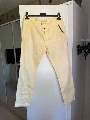 Gula chinos med raka ben - Snygga ljusgula chinos med raka ben och normal midja. Byxorna har två framfickor och dekorativa knappar vid ena fickan. Tillverkade i ett mjukt bomullstyg som känns skönt mot huden. Perfekta för dig som vill ha en färgklick i garderoben. Från märket Odd Molly i storlek 3 som enligt deras storlekguide motsvarar en storlek L. Helt nya med tyvärr är det något blekare vid låren se bild. 