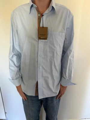 Ljusblå skjorta från Burberry - Säljer en ljusblå skjorta från Burberry med klassisk krage, bröstficka och broderad logga i modellen CLAVERDON. Skjortan har långa ärmar och knäppning framtill med Burberrys signaturrand på insidan av knappslån. Perfekt för en stilren look. Helt oanvänd, köpt i Milano. Nypris: 3400kr. Hör av dig om du har några frågor eller vill ha fler bilder! 