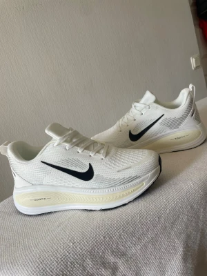 Nike - Snygga vita Nike sneakers med svart swoosh på sidan och en chunky sula med ZoomX-dämpning. Ovandelen är i mesh för bra andningsförmåga och sulan har svarta detaljer undertill. Perfekta för en sportig och clean look.