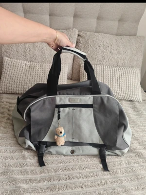 Grå weekendbag med nalle-nyckelring - Snygg och rymlig weekendbag i två nyanser av grått med svarta handtag och detaljer. Väskan har en praktisk ytterficka framtill och en söt liten nalle-nyckelring som dekoration. Tillverkad i slitstark textil, perfekt för resor eller gymmet.