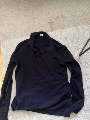 Mörkblå långärmad pikétröja - En stilren mörkblå långärmad pikétröja med klassisk krage och knappar framtill. Tröjan har en smal passform och är tillverkad i ett mjukt, tunt material som passar perfekt till olika outfits. Jag är storlek S 180 cm lång den är aning för liten för mig passar nog hur mycket du än väger väldigt töjbar