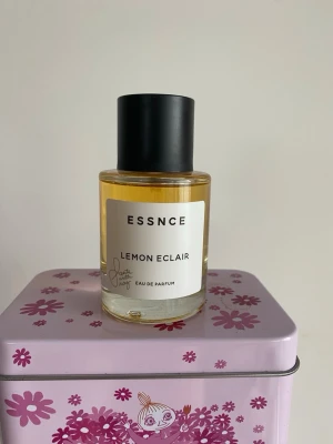 ESSNCE Lemon Eclair parfym - Fräsch parfym från ESSNCE med doften Lemon Eclair. Flaskan är cylindrisk i genomskinligt glas med svart lock och vit etikett. Innehållet är ljusgul vätska. Modern och stilren design som passar perfekt i badrumshyllan.