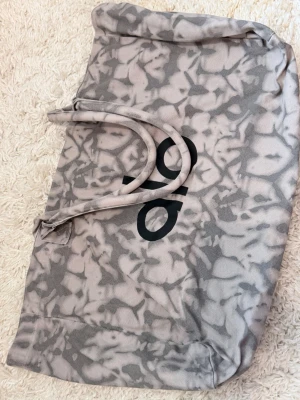 Alo Beige mönstrad weekendbag - Säljer en beige och gråmönstrad weekendbag med två handtag och tryckt svart logga. Väskan har mjuk form och är tillverkad i tygmaterial, perfekt för kortare resor eller gymmet.