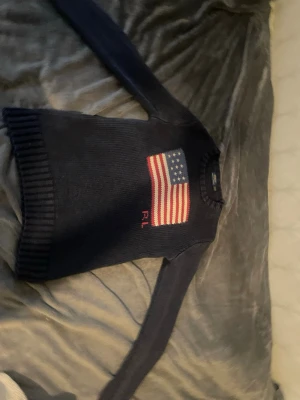 Mörkblå stickad tröja med USA-flagga - Snygg mörkblå stickad tröja med amerikansk flagga på bröstet och röda initialerna RL under flaggan. Tröjan har rund halsringning och ribbstickade muddar vid ärmslut och nederkant. Perfekt för dig som gillar klassisk och preppy stil. Lite urtvättad och tight vid armarna!!