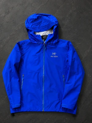 Arcteryx beta lt jacket  - Snygg blå regnjacka från Arc'teryx med huva och hel dragkedja framtill. Jackan har två sidofickor med dragkedja och är tillverkad i ett lätt, vattentätt material. Perfekt för regniga dagar och outdoor-äventyr.