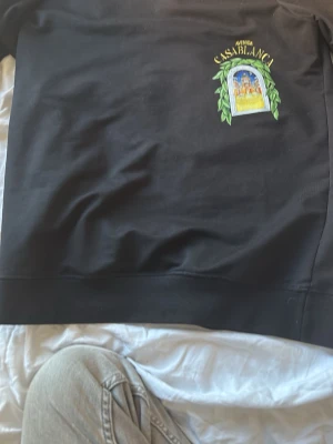 Svart sweatshirt från Casablanca - Säljer en svart sweatshirt från Casablanca med färgglatt tryck framtill. Trycket visar ett fönster med gröna blad runt och en byggnad i bakgrunden, samt texten 'CASABLANCA' i gult ovanför. Tröjan har rund hals och ribbade muddar. Perfekt för dig som gillar streetwear och snygga detaljer.