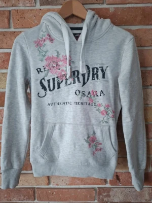Grå Superdry hoodie med blommor - Snygg ljusgrå hoodie från Superdry med tryckt logga och Osaka-text på bröstet. Hoodien har blommiga detaljer i rosa och grönt, huva med dragsko och en stor magficka. Perfekt för en avslappnad och trendig stil.