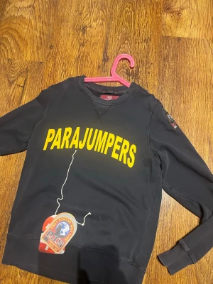 Parajumper tröja - Cool svart sweatshirt från Parajumpers med stor gul logga på bröstet och patch på ärmen. Klassisk rund halsringning och ribbade muddar. Perfekt för dig som gillar streetwear och vill ha något bekvämt och snyggt.