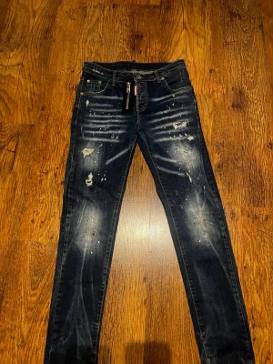 Dsquared2 jeans  - Säljer ett par mörkblå Dsquared2  jeans med slitningar och färgstänk för en trendig look. Jeansen har klassisk femficksmodell och dragkedja i gylfen. Perfekta för dig som gillar en edgy och avslappnad stil.