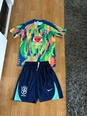 Brasilien Shorts Sett - Säljer ett Brasilien träningsset från Nike med färgglad t-shirt i grönt, gult, blått och rosa med Guaraná Antarctica-logga och CBF-märke. Matchande mörkblå shorts med Nike-logga och Brasilien-emblem. Perfekt för fotbollsträning eller sport.