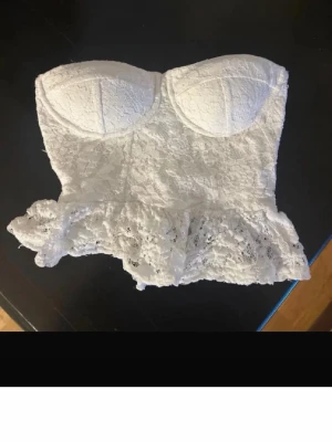 Vit spets med volang - Säljer en söt vit  i spets med vadderade kupor och volangkant nedtill. Bodyn har en romantisk känsla med sitt blommiga spetsmönster och figurnära passform. Perfekt för dig som gillar feminina detaljer och vill ha något extra under kläderna.