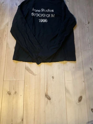 Svart långärmad tröja Acne Studios - Svart långärmad tröja från Acne Studios med tryck i vitt på bröstet där det står 'Acne Studios STOCKHOLM 1996'. Tröjan har en avslappnad passform och är perfekt för en cool och enkel stil.