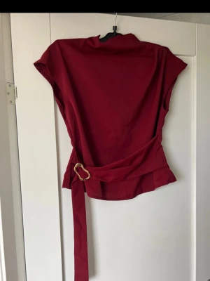 Vinröd topp med bälte och kort ärm - Snygg vinröd topp med korta ärmar och hög krage. Toppen har ett långt bälte i midjan med guldfärgat spänne som ger en cool detalj. Materialet ser ut att vara mjukt och lätt, perfekt för att styla med jeans eller kjol.
