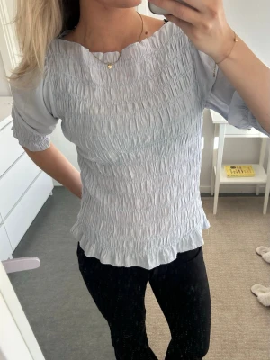 Ljusblå smockad blus med trekvartsärm - Supersöt ljusblå blus med smockad struktur och trekvartsärm. Blusen har en rund halsringning och lätt vågig kant nedtill. Perfekt att styla med både jeans och kjol för en trendig look.