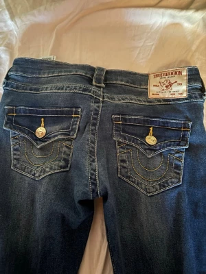 True Religion mörkblå bootcut jeans - Säljer ett par mörkblå bootcut jeans från True Religion med ikoniska bakfickor, kontrastsömmar och dekorativa knappar. Jeansen har en klassisk femficksdesign och patch med logga bak i midjan. Perfekta för dig som gillar en avslappnad men trendig stil. Endast använda en gång som nya storlek w24💕