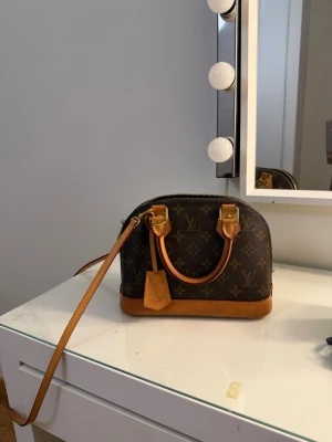 Louis Vuitton Monogram handväska - Säljer min fina Louis Vuitton Alma BB i Monogram Canvas, köpt 2022.  Väskan är använd men väl omhändertagen och är överlag i fint begagnat skick. Det finns visst normalt slitage på läderdetaljerna efter användning, vilket syns på bilderna. Monogram-canvasen är i fint skick.  Originalkvitto och originalbox medföljer. Väskan är äkta från Louis Vuitton.