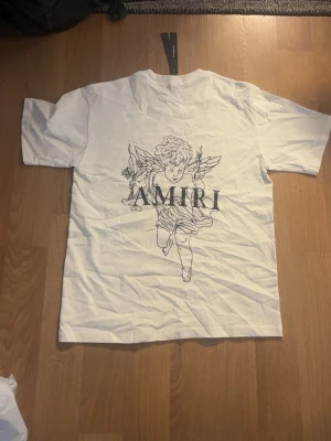 Vit AMIRI t-shirt med ängeltryck - Säljer en vit t-shirt från AMIRI med ett stort tryck av en ängel och logga på framsidan samt mindre logga med ängel på bröstet. Klassisk passform och rund hals. Perfekt för dig som gillar streetwear och stilrena prints. Strl L men sitter som en M