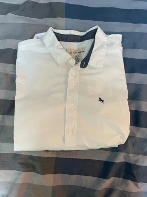 Vit skjorta med broderad detalj - Klassisk vit skjorta med knappar framtill och en liten broderad figur på bröstet. Skjortan har en krage med kontrastfärg på insidan och är perfekt för en clean och stilren look. Tillverkad i bomull för skön känsla.