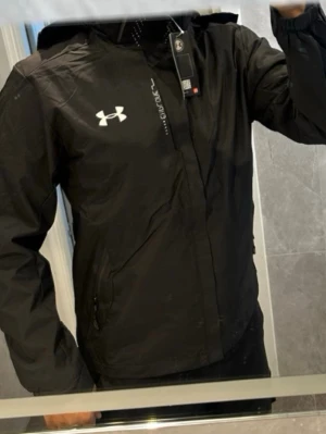 Svart vindjacka från Under Armour - Snygg svart vindjacka från Under Armour med huva och vita logodetaljer på bröstet. Jackan har dragkedja framtill, två sidofickor med dragkedja och elastiska muddar vid ärmsluten. Perfekt för blåsiga dagar och sportiga outfits.