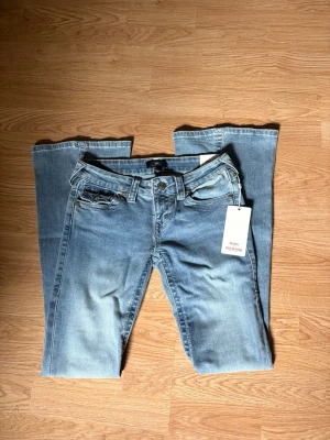 True Religion Joey flare jeans blå - Snygga blå jeans från True Religion, modell Joey med low rise och flare-ben. Jeansen har slitna detaljer och klassisk femficksdesign. Perfekta för dig som gillar en avslappnad men trendig look.