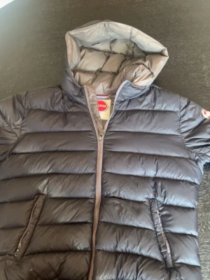 Mörkgrå dunjacka från Colmar - Snygg mörkgrå dunjacka från Colmar med huva och två dragkedjeförsedda fickor. Jackan har quiltad design och hög krage för extra värme. Perfekt för kalla vinterdagar och riktigt skön att bära.