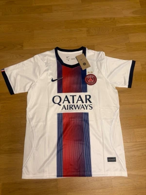 Kvaratskhelia borta 26/27 M - Snygg PSG matchtröja i vitt med vertikala ränder i rött och blått. Tröjan har korta ärmar, svart krage och muddar samt PSG-logga och Nike-logga på bröstet. Qatar Airways sponsortryck på framsidan och Kvaratskhelia 7 på ryggen.
