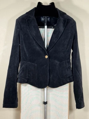 Armani Jeans Blazer / Dam EU 38–40 (S-M) – Svart Velour / Enknapps AJ-knapp - Storlek enligt märkning: [KONTROLL] Passar: S–M / EU 38–40  Mått (plagget liggande platt): Bröstmått (arm till arm): 42 cm Totallängd: 54 cm Ärmlängd: 66 cm  Armani Jeans kort cropped blazer/jacka i svart syntetisk velour med fint ribbmönster. Modellen har enknappsknäppning med guldtonad AJ-logotypknapp, klassiska notch-lappar, två påsydda patch-fickor framtill samt helfodringsinteriör i polyester.  Mycket gott begagnat skick – blazern är mörk, ren och välvårdad. 0105