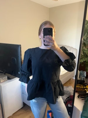 Black Puff Sleeve Peplum Top with Bow Back Detail - This is a black long-sleeve blouse featuring puffed sleeves and a peplum hem. The back of the blouse has a stylish open design with two bow tie closures—one at the upper back and one at the mid-back, creating a chic and feminine look. The fabric appears to be lightweight with a slight sheen, giving it a dressy yet playful vibe. The top is paired with light-wash jeans for a casual yet trendy outfit.