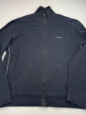 Armani Jeans Mörkblå zip hoodie med fickor - Mörkblå hoodie från Armani Jeans  med dragkedja framtill och två stora magfickor. Modellen har hög krage och ribbade muddar vid ärmslut och nederkant. Liten diskret logga på bröstet. Perfekt för en avslappnad och stilren look.0097