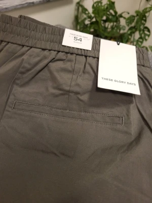 Olivgröna shorts från These Glory Days - Snygga olivgröna shorts från These Glory Days med elastisk midja och knappstängning. Shortsen har raka ben, sidofickor och en bakficka. Perfekta för varma dagar och har en clean, minimalistisk look.